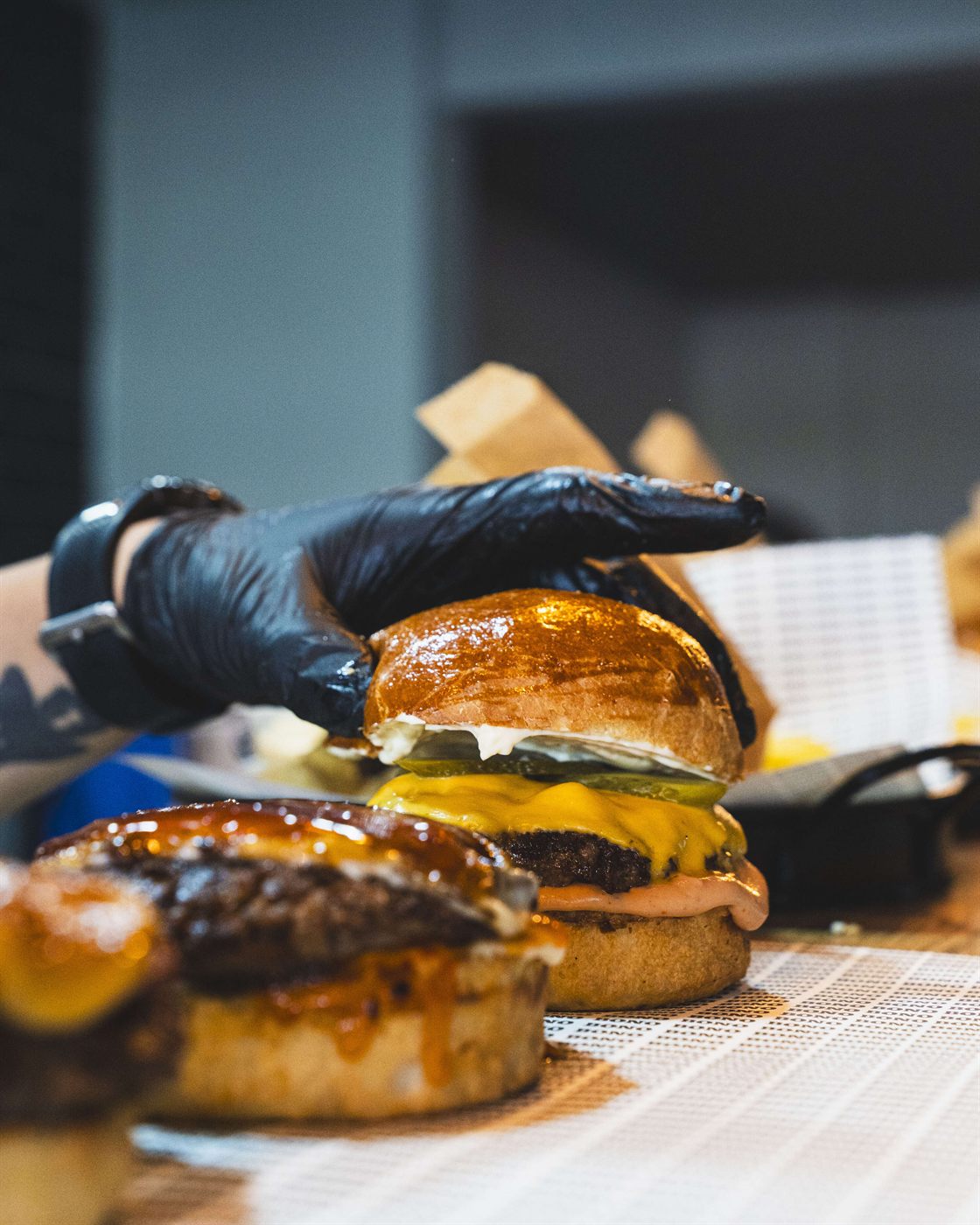 Siyah eldivenli bir el, cheddarlı burgerin üzerine parlak ekmeği yerleştiriyor